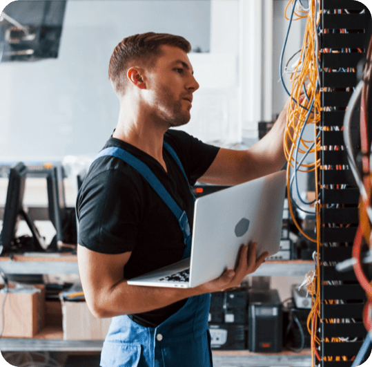 A man in overalls stands holding a laptop in front of a rack filled with various wires and cables.