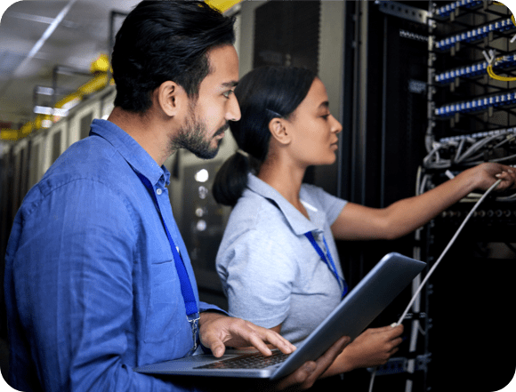 Two technicians collaborating on a server inside a data center, surrounded by rows of equipment and blinking lights.